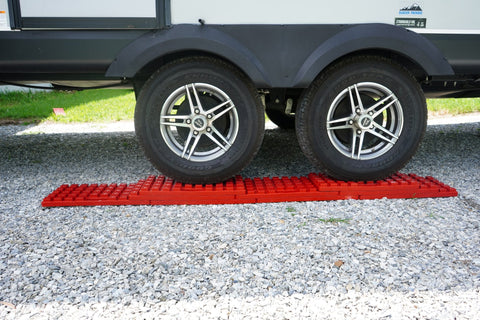 Red interlocking leveling blocks laid out in front of the tandem axle tires of a travel trailer on a gravel campsite.