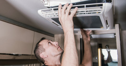  A man taking the cover off the main vent of his RV air conditioning unit. He is holding the cover with both hands.